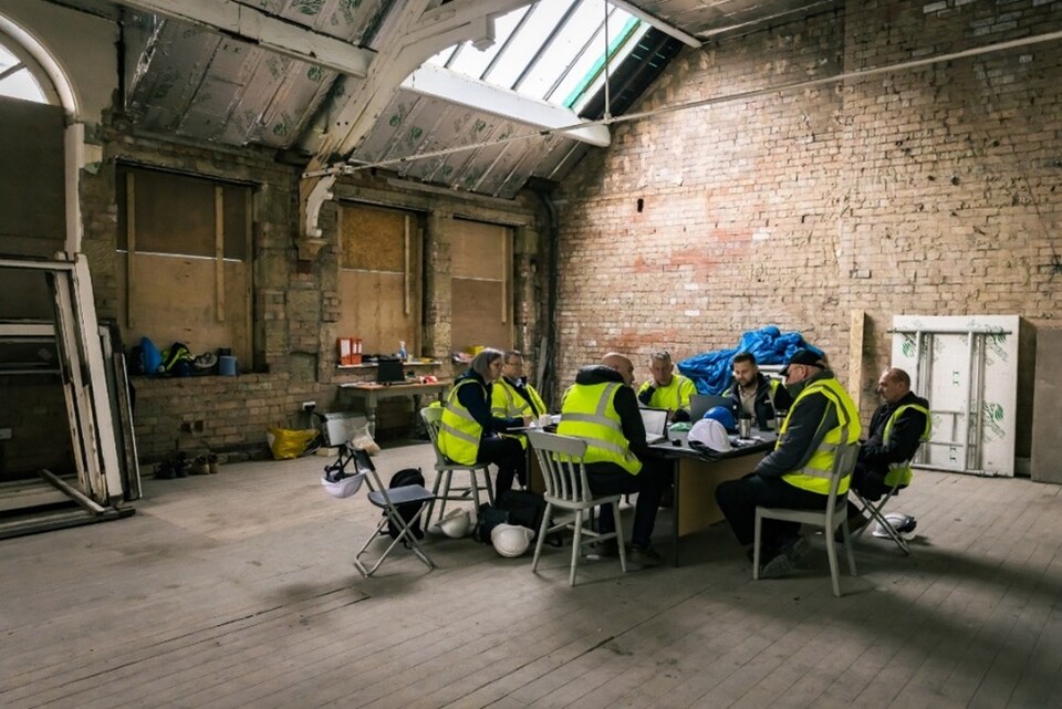 A group of people wearing yellow safety vests sit around a table in a large, unfinished room with exposed brick walls, wooden floors, and a partially covered ceiling. Construction materials are scattered around.